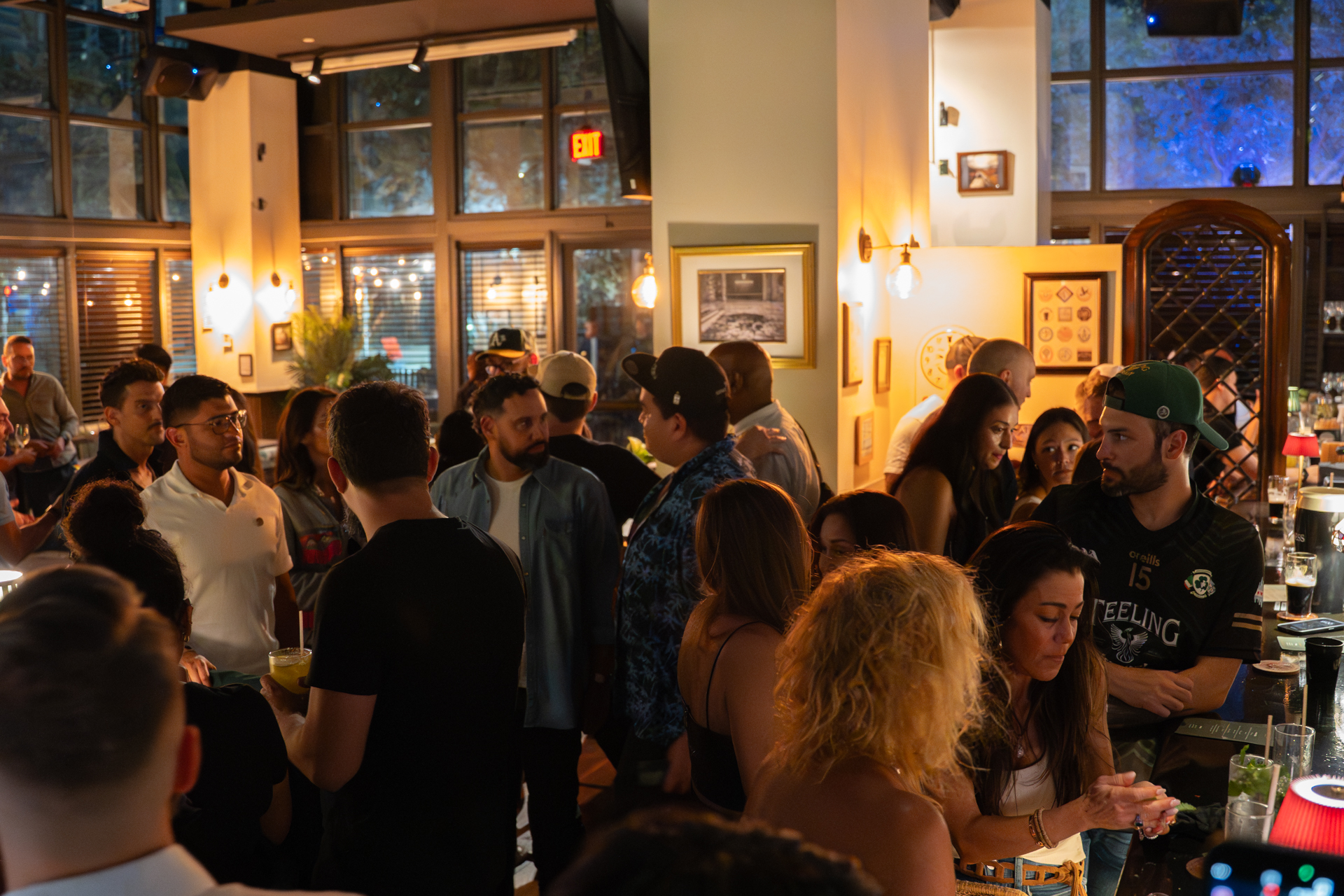 A lively bar scene with people socializing, standing, and talking. The space is warmly lit with large windows, framed art on the walls, and various drinks on the bar counter. South Florida Business & Wealth
