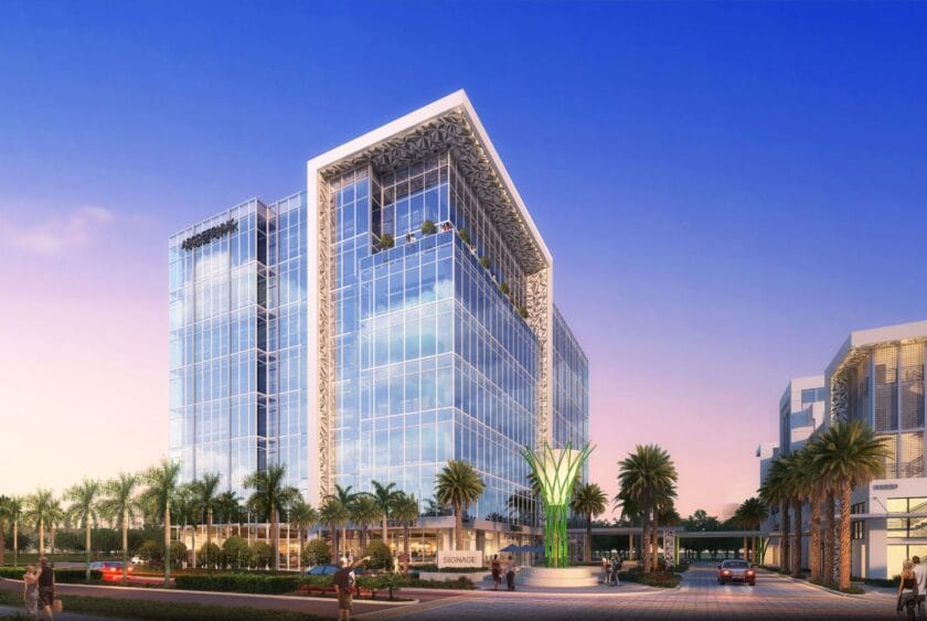Modern glass office building with palm trees in front, people walking nearby, and a decorative green sculpture at the entrance, under a clear blue and pink sky at sunset. South Florida Business & Wealth