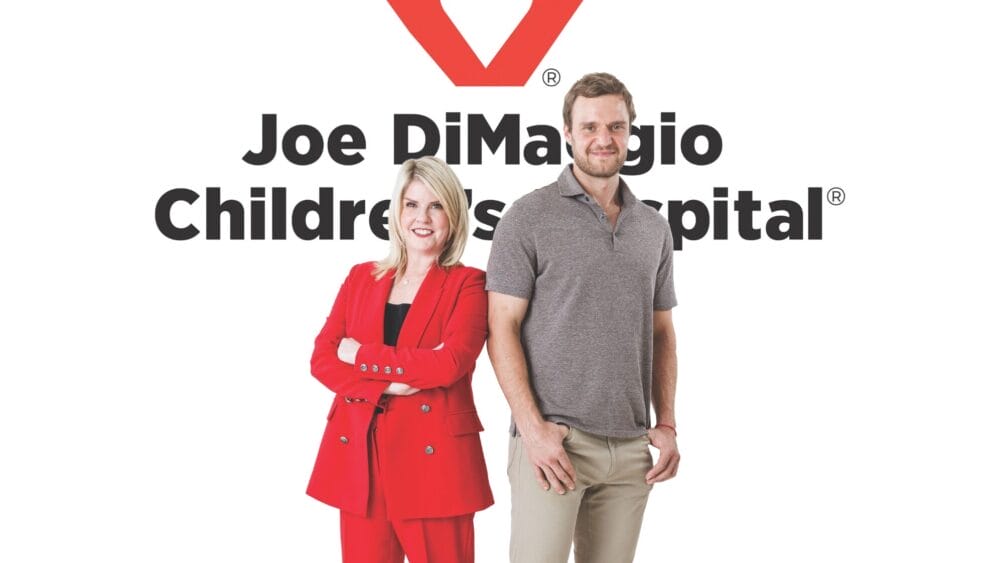 Two adults stand in front of a sign reading "Joe DiMaggio Children’s Hospital." The woman on the left wears a red suit and smiles with arms crossed. The man on the right wears a gray polo shirt and khaki pants, smiling with a hand in his pocket. South Florida Business & Wealth