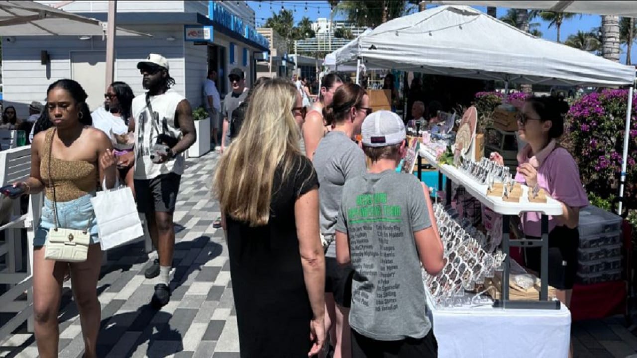 People browse various booths at an outdoor market on a sunny day. Some are shopping, others are looking at jewelry displays, and palm trees and tents are visible in the background. South Florida Business & Wealth