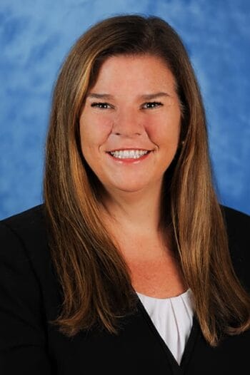 A woman with long brown hair wearing a black blazer and white top smiles at the camera, posed against a blue background. South Florida Business & Wealth