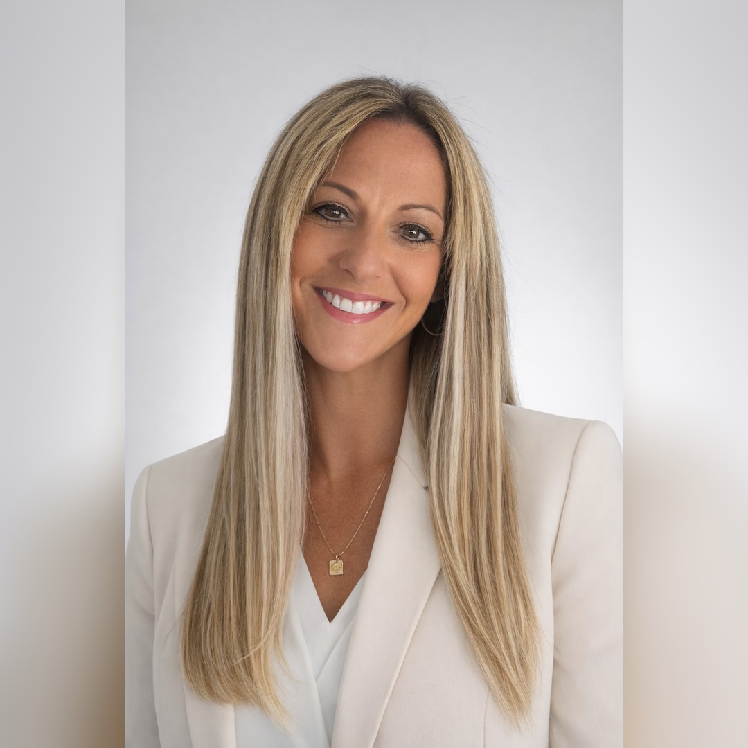 A woman with long straight blonde hair, wearing a light-colored blazer and white top, smiles at the camera against a plain, light gray background. She is wearing a gold necklace and hoop earrings. South Florida Business & Wealth