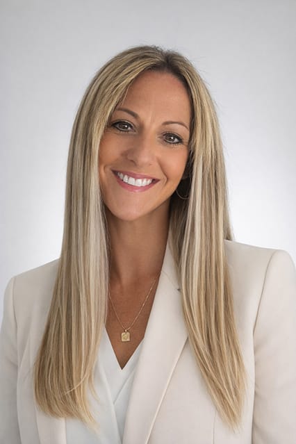 A woman with long straight blonde hair, wearing a white blazer over a light blouse, smiles at the camera against a plain light background. She wears a gold necklace and hoop earrings. South Florida Business & Wealth