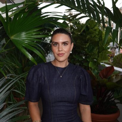 A woman with dark hair tied back, wearing a dark denim dress, stands among lush green plants on a balcony. She has natural makeup, pearl earrings, and a calm expression. The background shows more greenery and part of a building. South Florida Business & Wealth