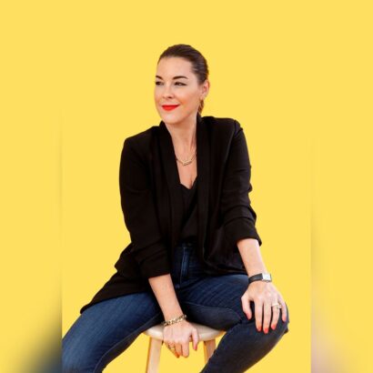 A woman with dark hair tied back, wearing a black blazer, jeans, and gold jewelry sits on a stool against a bright yellow background, smiling and looking to her left. South Florida Business & Wealth