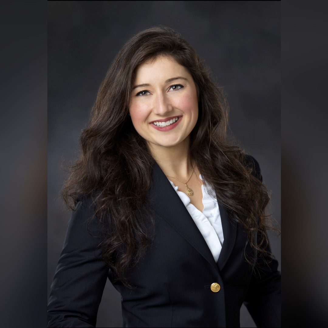 A woman with long wavy brown hair, wearing a dark blazer over a white blouse, smiles at the camera against a dark, blurred background. South Florida Business & Wealth