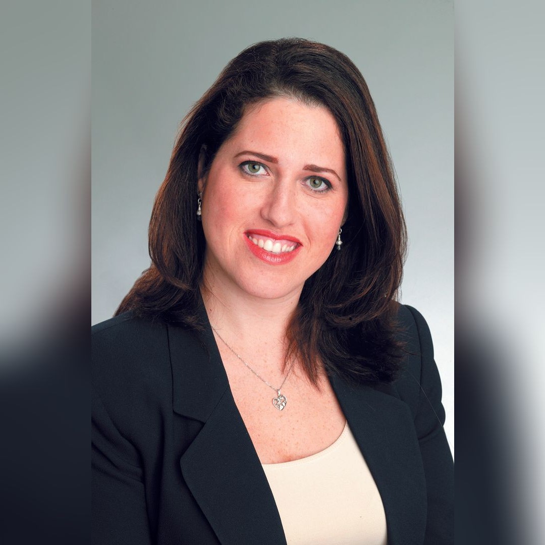 A woman with long brown hair, wearing a black blazer over a light top, smiles at the camera. She has light skin, is wearing earrings and a heart-shaped necklace, and is posed against a plain gray background. South Florida Business & Wealth