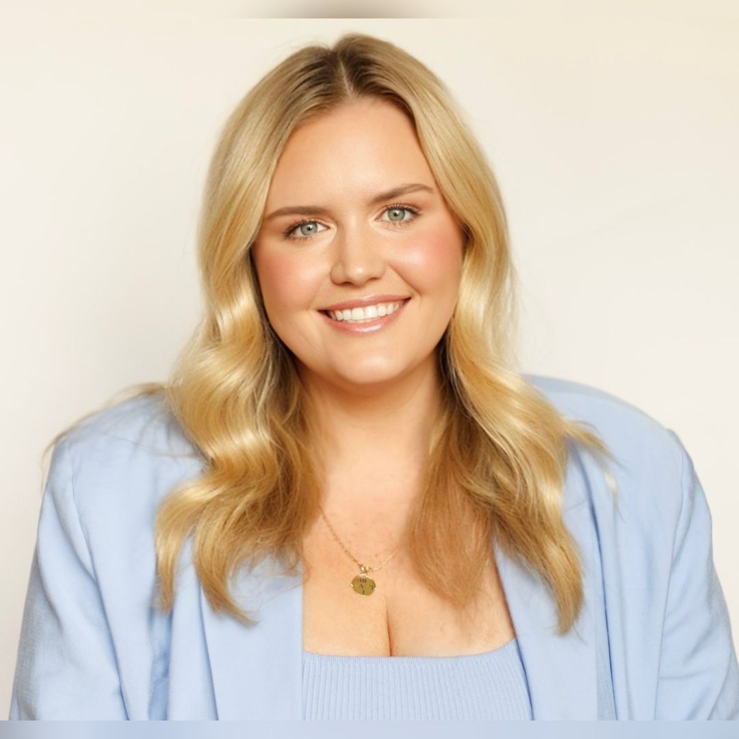 A woman with long blonde hair smiles at the camera. She is wearing a light blue blazer over a matching top and a gold necklace with a round pendant. The background is plain and light-colored. South Florida Business & Wealth