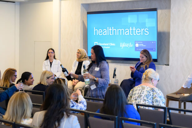 A woman holding a microphone stands among seated attendees at a panel discussion, with four women panelists at the front. A screen behind them displays "healthmatters Cleveland Clinic". South Florida Business & Wealth