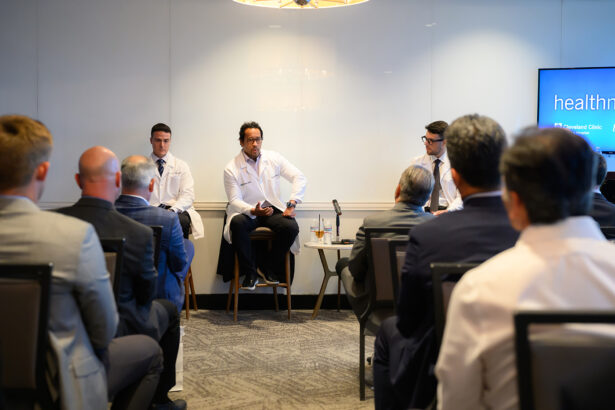 Three men in white coats sit at the front of a conference room, speaking to an audience of seated attendees. A screen to the right displays the beginning of the word "health." The setting appears professional and formal. South Florida Business & Wealth