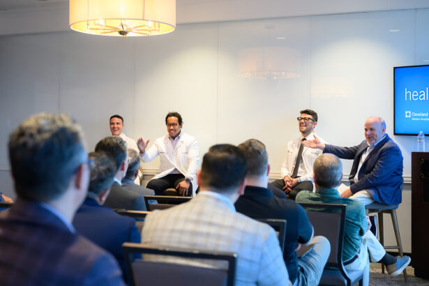 A group of four men sit at the front of a room, engaging in a panel discussion with an audience. Two men wear white medical coats; the others wear suits. One speaker gestures while the audience listens attentively. South Florida Business & Wealth