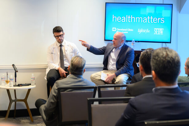 A man in a suit gestures while speaking to a man in a white coat on a panel in front of an audience. A screen behind them displays "healthmatters" and logos for Cleveland Clinic, Lifestyle, and SBW. South Florida Business & Wealth