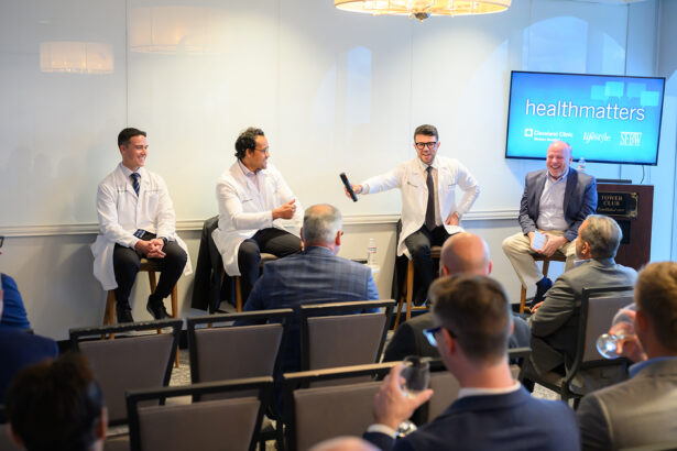 Four men, three in white lab coats and one in a suit, sit on stools at the front of a room addressing an audience. A screen behind them displays “healthmatters” and several logos. The audience listens attentively. South Florida Business & Wealth