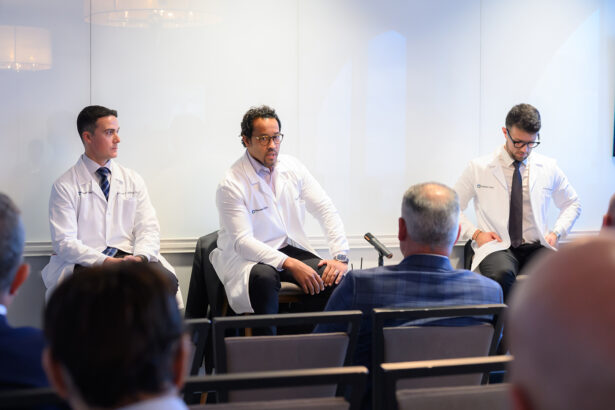Three doctors in white coats sit at the front of a room, facing an audience during a discussion or panel event. Two audience members are visible from behind in the foreground. South Florida Business & Wealth