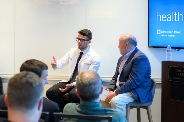 A doctor in a white coat speaks and gestures with his hand while seated next to another man in a suit during a panel discussion in front of an audience. A screen displays the Cleveland Clinic logo in the background. South Florida Business & Wealth