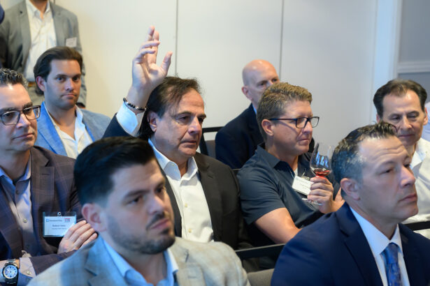 A group of men in business attire sit in a conference room. One man in the center raises his hand, while others listen attentively. Some wear name badges and hold drinks. The atmosphere appears professional and focused. South Florida Business & Wealth