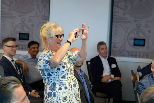 A woman in a floral dress stands and smiles while taking a photo with her phone at an indoor event. Several people in business attire are seated around her, watching and smiling. South Florida Business & Wealth