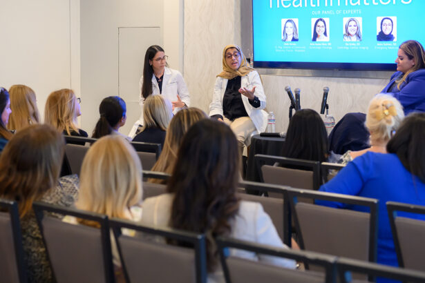 A diverse panel of female health experts speaks to an audience in a conference room. One panelist gestures while others listen. The audience, mostly women, faces the stage and presentation screen reading "health matters. South Florida Business & Wealth