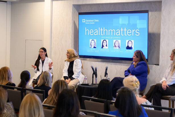 Four women sit on a panel in front of an audience, discussing health topics at a Cleveland Clinic event. A screen behind them reads "healthmatters" and displays their names and photos. Two microphones are set up in front of the panel. South Florida Business & Wealth