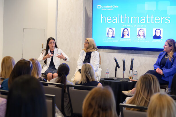 A panel of female medical experts sits in front of an audience at a Cleveland Clinic "healthmatters" event. One expert gestures while speaking, while others listen attentively. A screen behind displays panelist names and photos. South Florida Business & Wealth