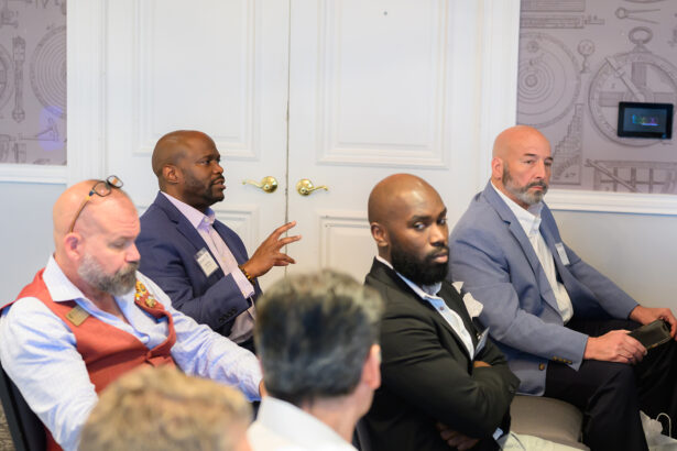 A group of men in business attire sit attentively in a conference room; one man in a blue blazer is speaking and gesturing with his hand while others listen. South Florida Business & Wealth