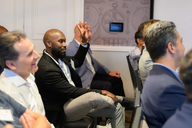 A man in a black blazer and plaid pants sits among others in a conference room, raising his hand to speak or ask a question. People around him are listening attentively, and the atmosphere appears professional. South Florida Business & Wealth