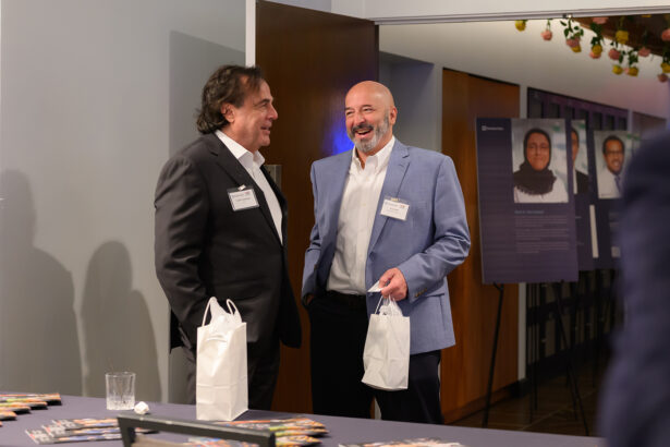 Two men in business attire stand and chat at an indoor event, each holding a white gift bag. Behind them are informational posters displayed on easels and a table with more bags and pamphlets in the foreground. South Florida Business & Wealth