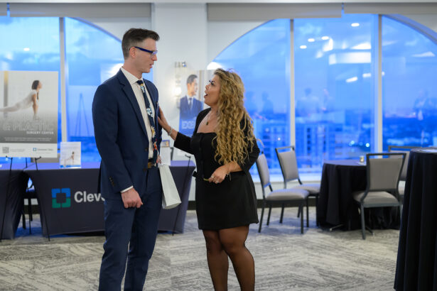 A man in a suit and a woman in a black dress stand talking in a modern conference room with large windows and city views. Event banners and empty chairs are visible in the background. South Florida Business & Wealth