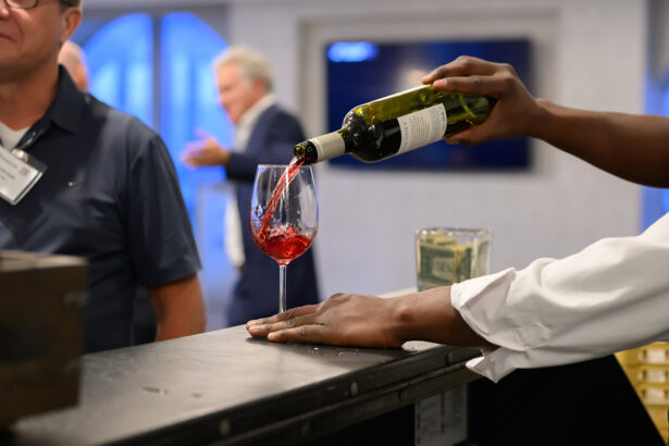 A person pours red wine into a glass at a bar, with a cash tip jar nearby. People are blurred in the background, suggesting a social or networking event. South Florida Business & Wealth