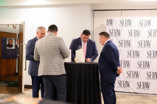 Four men in suits stand around a tall black table, engaged in conversation at an indoor event. A step-and-repeat backdrop with "SFBW" branding is visible behind them. South Florida Business & Wealth