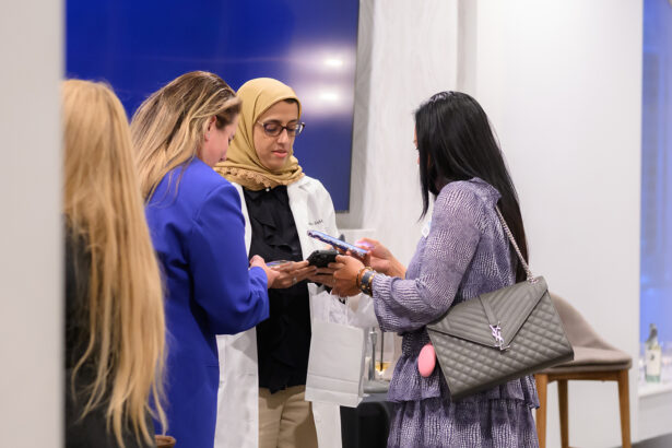 Three women stand together indoors, looking at their phones. One woman wears a white lab coat and a tan hijab, while the others are dressed in business attire. They appear to be engaged in conversation or sharing information. South Florida Business & Wealth