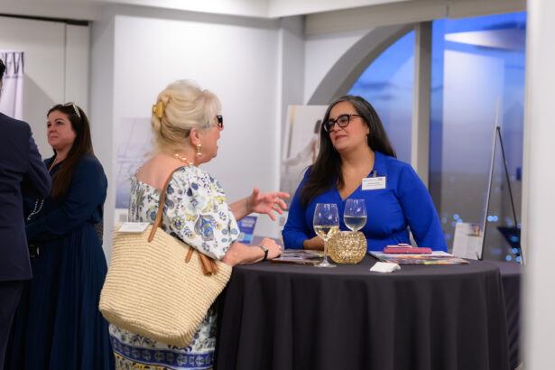 Two women converse at a round table with drinks and brochures during an indoor event. One woman wears a blue dress and glasses, the other a floral dress and straw bag. Another person stands in the background. South Florida Business & Wealth