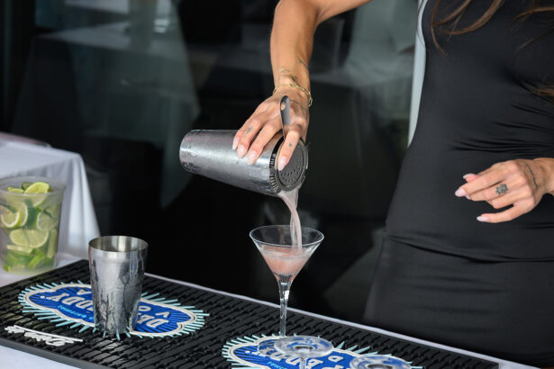 A person in a black and gray dress pours a pink cocktail from a metal shaker into a martini glass at a bar, with lime wedges visible on the side. South Florida Business & Wealth