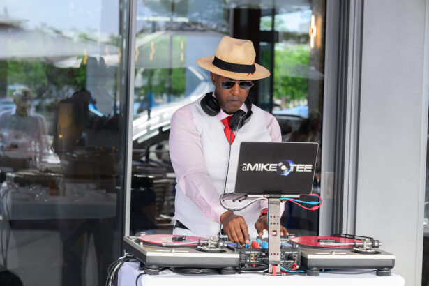 A DJ wearing sunglasses, a straw hat, and a white vest with a red tie is mixing music on turntables outdoors. Headphones rest around his neck, and his laptop is open on the stand in front of him. South Florida Business & Wealth