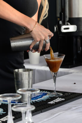 A person pours a brown cocktail from a shaker into a martini glass on a bar counter, with empty glasses and bar tools in the foreground. South Florida Business & Wealth