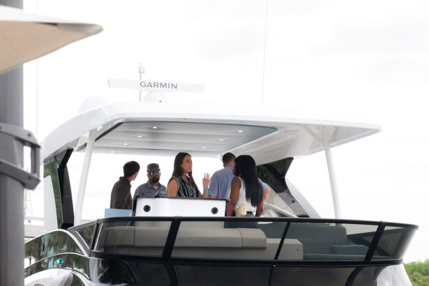 A group of people socializes on the deck of a modern yacht with a white roof and black railings, docked near the shore under a cloudy sky. The yacht displays a Garmin navigation system. South Florida Business & Wealth
