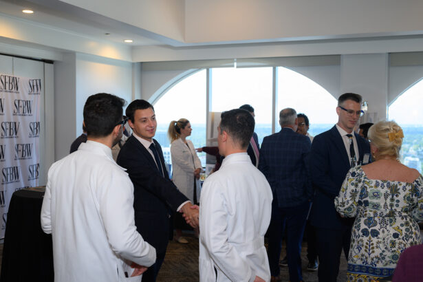 A group of people in business and medical attire are networking indoors; two men in focus are shaking hands, while others engage in conversation. Large windows in the background let in daylight. South Florida Business & Wealth