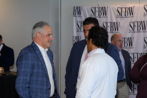 A group of men in business attire converse at a networking event with an SFBW (South Florida Business & Wealth) banner in the background. South Florida Business & Wealth