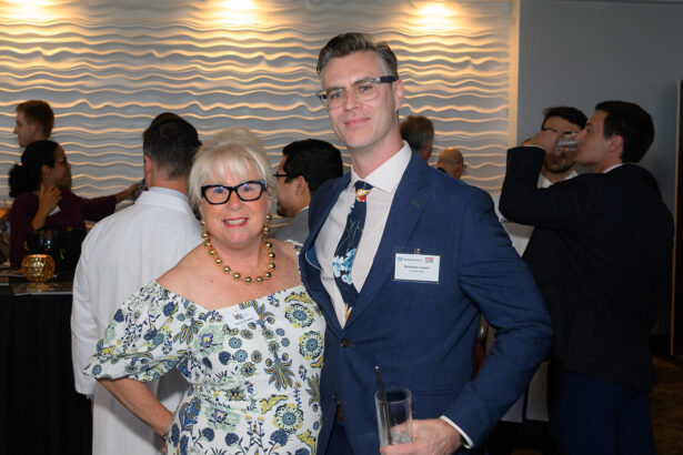 A woman and a man, both wearing glasses and floral-patterned clothing, smile and pose together at a social event. Other people mingle in the background in a room with a wavy textured wall. South Florida Business & Wealth