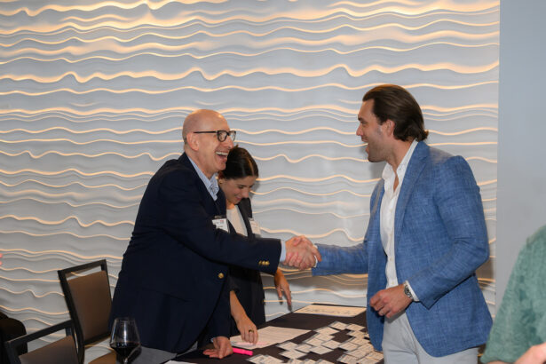 Two men shake hands and smile at a registration table, while a woman organizes nametags in the background; the setting features a modern, wavy-textured wall. South Florida Business & Wealth