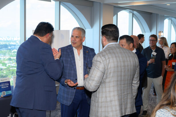 A group of professionally dressed men stand and converse at a networking event in a bright room with large windows. Other attendees are mingling in the background. South Florida Business & Wealth
