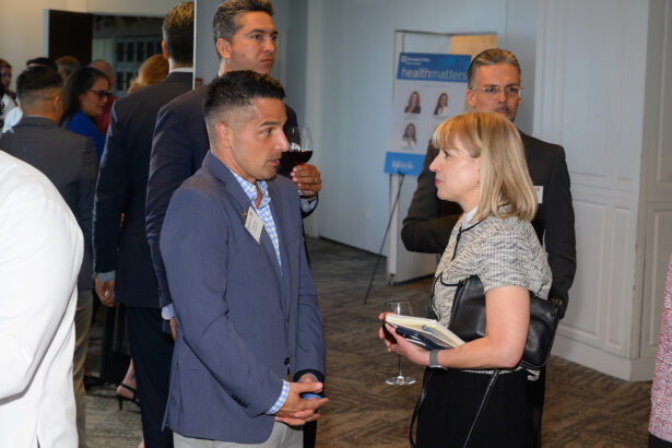 A group of professionally dressed people are socializing at an indoor networking event. A man and woman in the foreground are engaged in conversation, each holding a drink. Others are mingling in the background. South Florida Business & Wealth