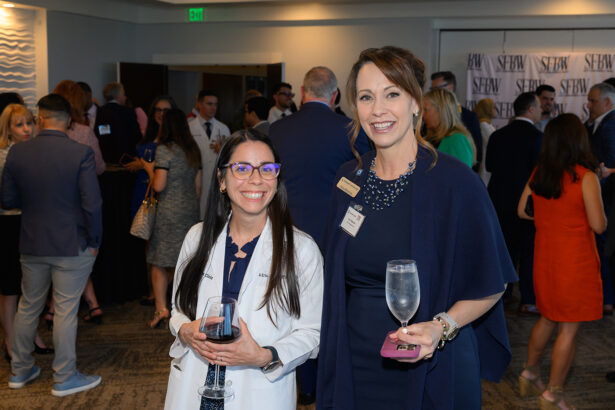 Two women stand smiling at a business networking event. One holds a glass of wine, the other a glass of water. People in business attire are conversing in the background, with an SEBW banner visible. South Florida Business & Wealth