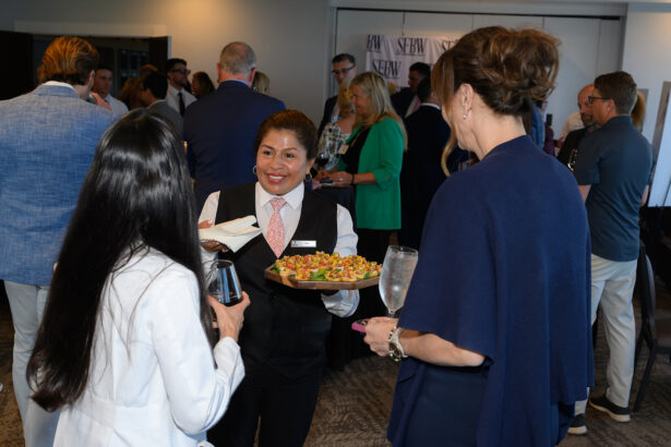 A server in a white shirt and pink tie offers appetizers to two women at a busy indoor networking event. People in business attire are mingling and talking in the background. South Florida Business & Wealth