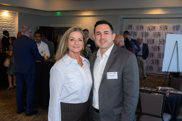 A woman and a man stand together, smiling, at a business networking event. Both are wearing business attire with name tags. Other attendees mingle in the background near tables, and SEBW banners are displayed. South Florida Business & Wealth