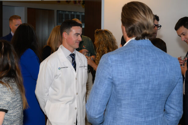 A man wearing a white lab coat stands among a group of people in business attire, engaging in conversation at a social or professional event indoors. South Florida Business & Wealth