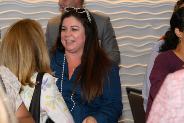 A woman with long dark hair wearing sunglasses on her head and a blue top smiles and talks with another person at an indoor event. Other people mingle in the background in front of a wavy-textured wall. South Florida Business & Wealth