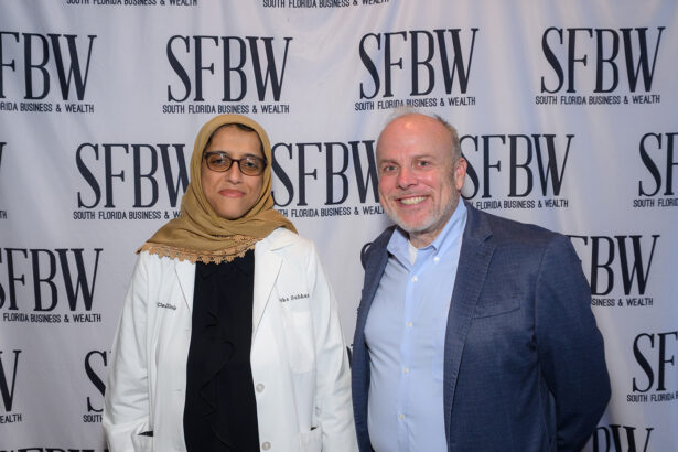 Two people pose for a photo in front of a backdrop with the text "SFBW South Florida Business & Wealth." One wears a tan hijab and white coat, and the other wears a light blue shirt and blazer, both smiling at the camera. South Florida Business & Wealth