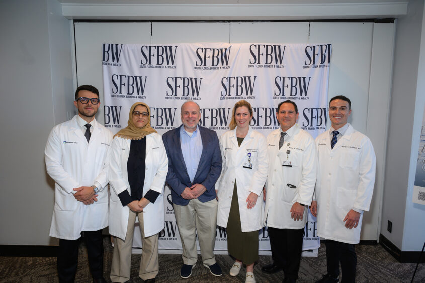 Six people, five in white lab coats and one in a blue blazer, stand smiling in front of a backdrop with repeated "SFBW" logos at an indoor event. South Florida Business & Wealth