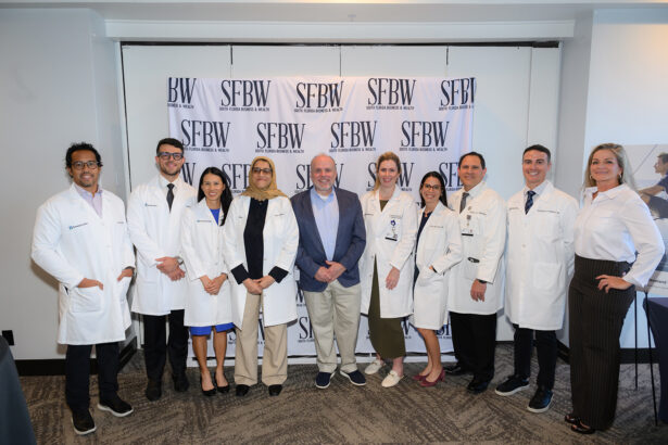 A group of ten people, most wearing white lab coats, stand together and smile for a photo in front of an SFBW (South Florida Business & Wealth) step-and-repeat banner in a conference room. South Florida Business & Wealth